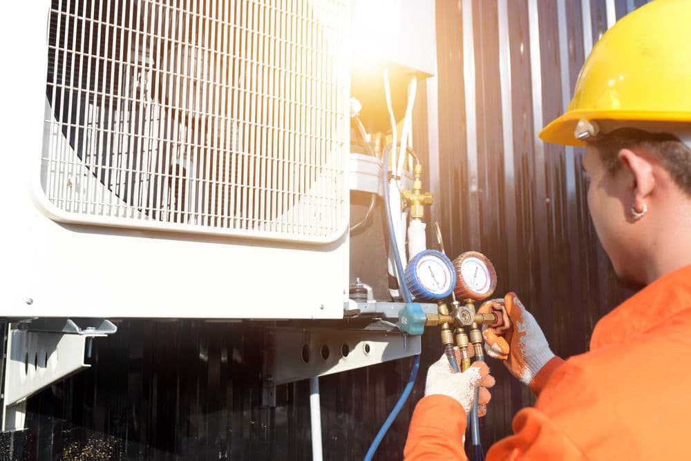 Technician checking HVAC system with gauges, wearing orange uniform and helmet, outdoor setting.